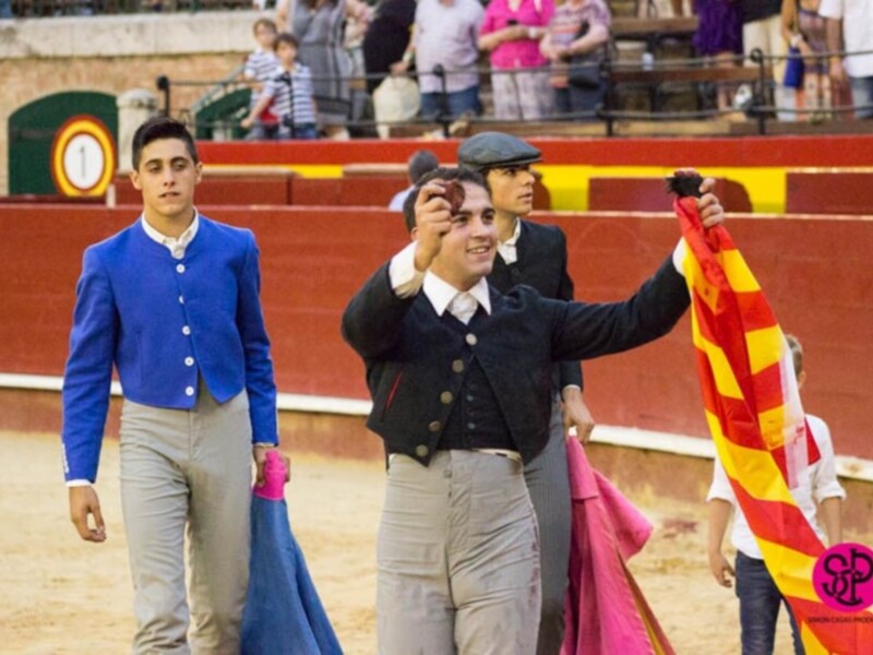 Jóvenes participantes en una corrida de toros, uno sosteniendo una bandera roja y amarilla.
