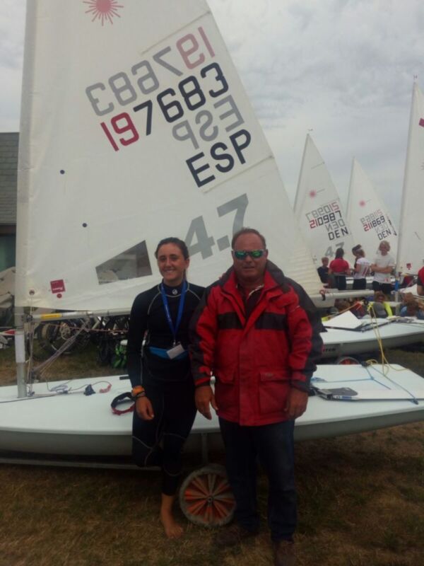Dos personas posan junto a una vela de barco de vela con las iniciales 'ESP' y '4.7', en un evento deportivo al aire libre.