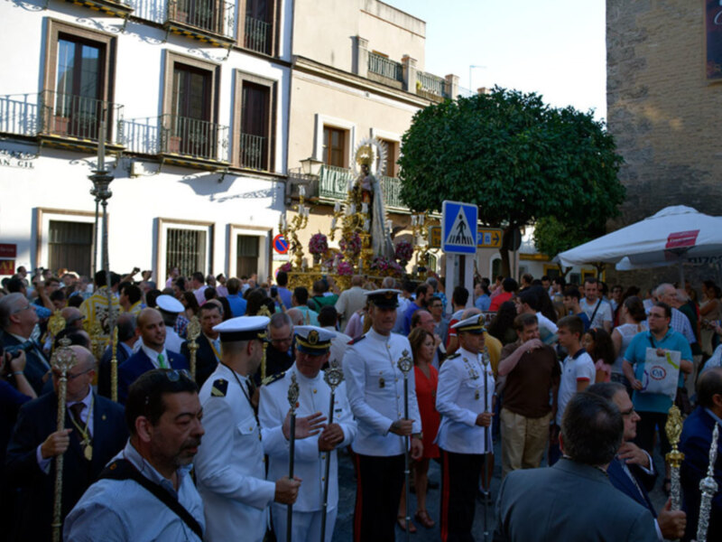 Fotografía de una procesión religiosa en una calle con edificios blancos y azules, gente vestida con uniformes militares y una figura venerada en el centro.