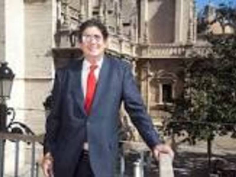 Man wearing a blue suit and red tie standing in front of a building with a tree in the background.