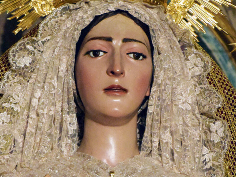 Imaginaria de una Virgen María vestida con una vestimenta tradicional y una aureola dorada, con un fondo que sugiere una iglesia o templo.
