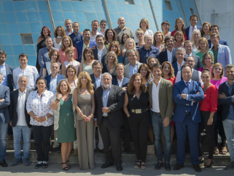Grande grupo de personas posando juntos en una escalinata al aire libre, frente a un edificio moderno con azulejos azules.