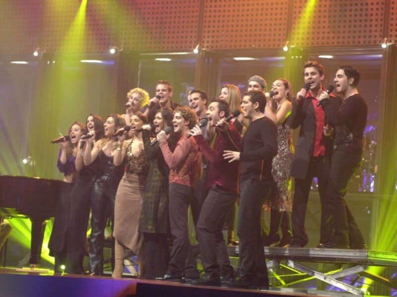 Grupo de cantantes en escenario con luces de colores, actuando con energía.