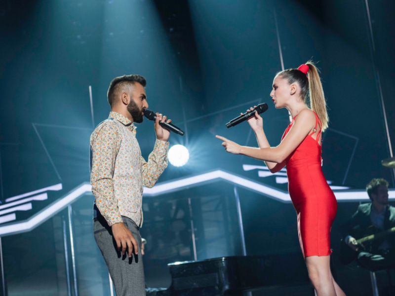 Cantantes en escenario con luces dramáticas y un músico tocando en el fondo.