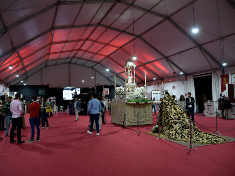 Una gran carpa con techo de tela roja, iluminada por luces blancas. Dentro hay una mesa grande con flores y velas, rodeada de personas observando. La carpa está en un espacio abierto con una alfombra roja y varios asientos de plástico.