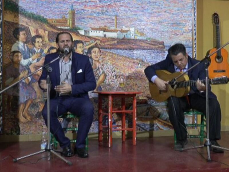Dos músicos interpretando en un escenario con un fondo artístico de paisaje urbano.