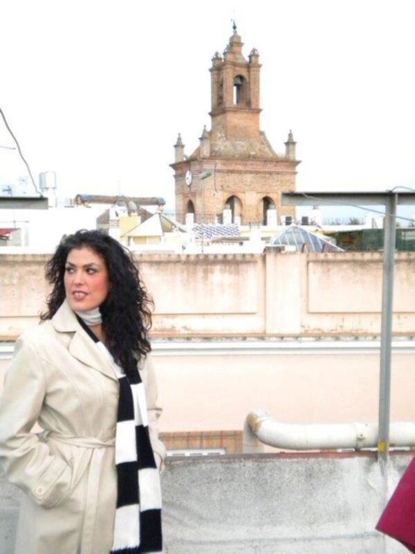 Mujer con abrigo beige y bufanda a cuadros en un entorno urbano con torre de iglesia al fondo.