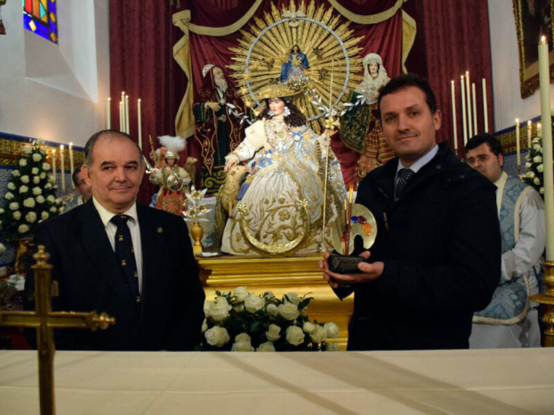 Celebración religiosa en una iglesia con dos hombres en trajes formales y una imagen de la Virgen María en el altar.