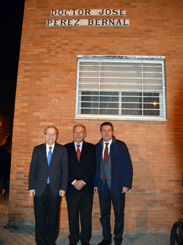 Se observa a tres hombres vestidos con trajes de negocios, posando frente a una pared de ladrillos rojos. En la parte superior de la pared, se puede leer el nombre "DOCTOR JOSE PEREZ BERNAL". La escena parece ser nocturna, con la luz de una ventana en el edificio iluminando los rostros de los hombres.