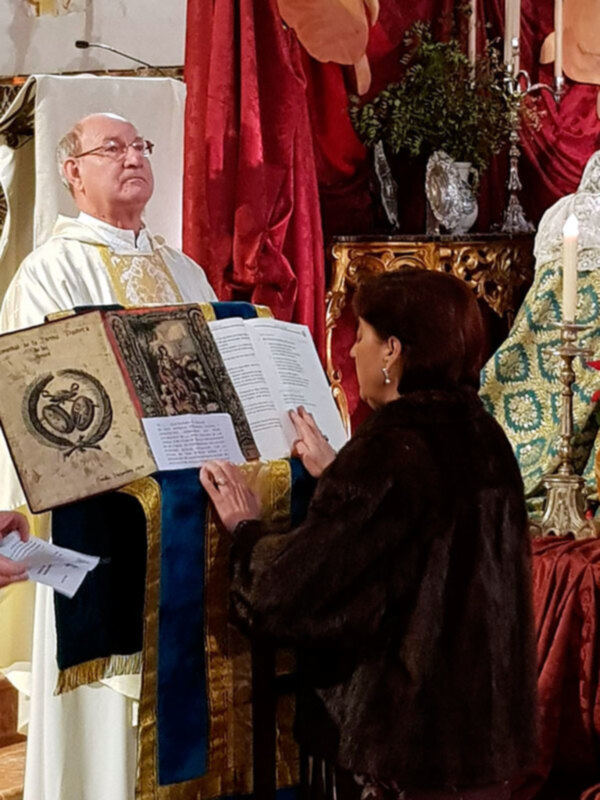 Una persona vestida con un manto negro y una corona de flores, sosteniendo un libro con un diseño antiguo, en un entorno ceremonial con velas y velos rojos.