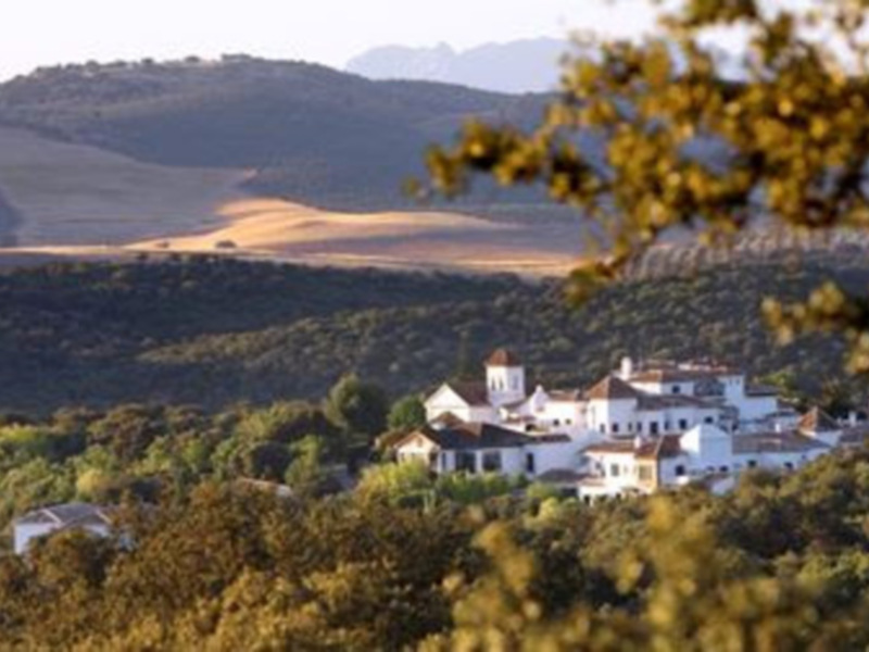 Vista panorámica de una pequeña aldea en el valle, rodeada de colinas y árboles en otoño, con un cielo claro y un horizonte visible.