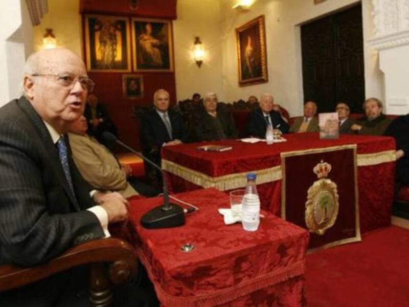 Un hombre en traje hablando en una sala formal con otros asistentes sentados en el fondo.