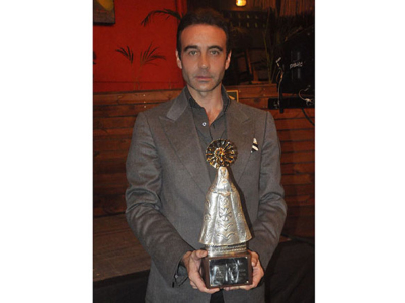 Persona con traje gris sosteniendo un trofeo con diseño ornamental en un ambiente interior con pared de madera y plantas.