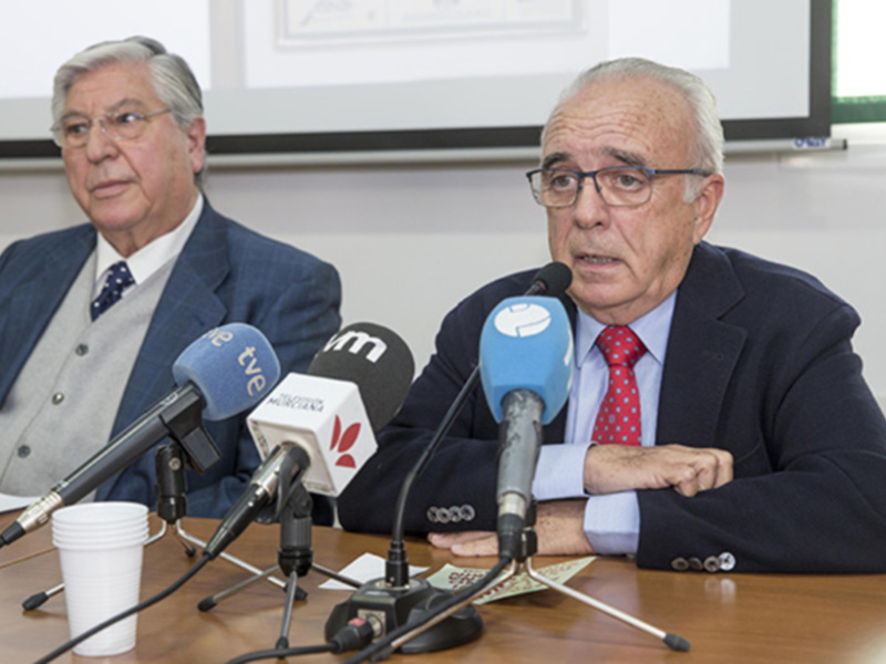 Dos hombres en traje sentados detrás de un escritorio con micrófonos, probablemente en una conferencia de prensa o reunión oficial.