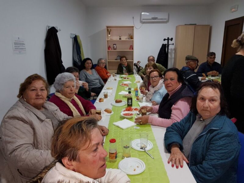 Una grupo de personas sentadas en una mesa durante un evento social, posiblemente una reunión o celebración. La mesa está cubierta con mantel y hay varios platos, vasos y botellas de bebidas. La sala tiene estanterías con libros y objetos decorativos, y una puerta visible en el fondo. La gente parece estar disfrutando de la reunión, con algunas personas sonriendo o hablando.