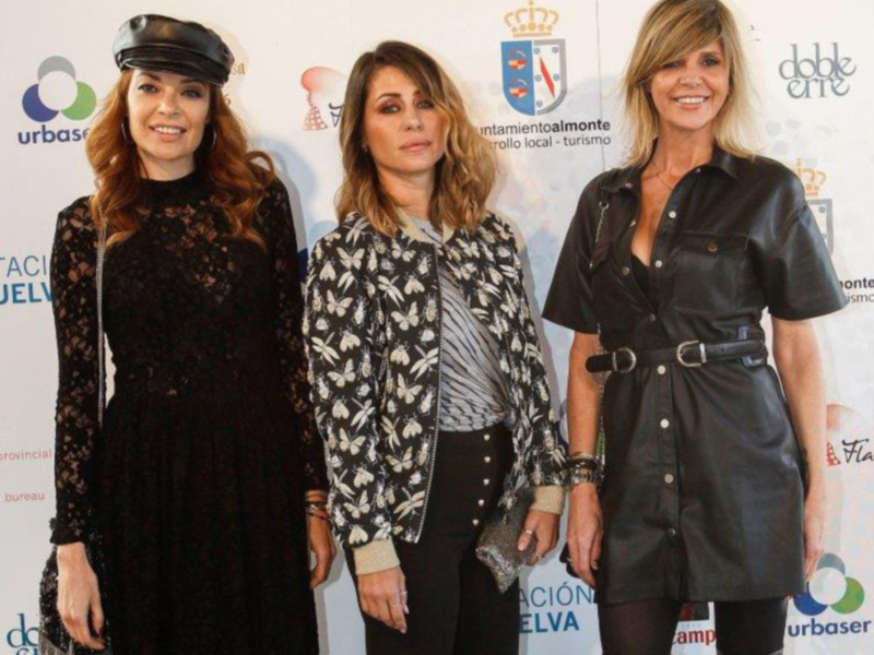 Tres mujeres posando juntas en un evento formal con fondos de patrocinadores visibles.