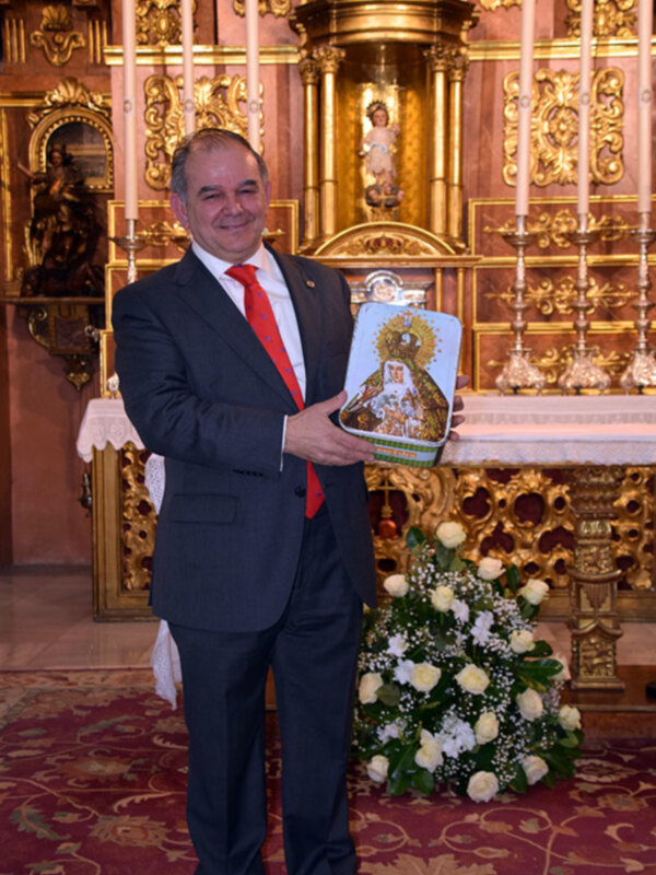 Hombre en traje con corbata roja y corbata de punta, sosteniendo un libro en un altar con detalles dorados y velas.