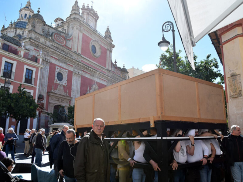 Procesión en la plaza histórica con edificios barrocos y una plataforma con personas en el centro.