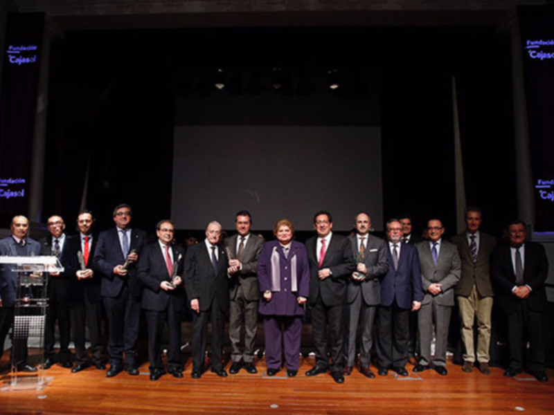 Una imagen de una ceremonia de premiación donde varios hombres y una mujer se encuentran en un escenario, todos sonrientes y algunos sostienen trofeos. La iluminación es suave, y el fondo muestra un gran pantalla con la inscripción 'Fundación Cajasol'.