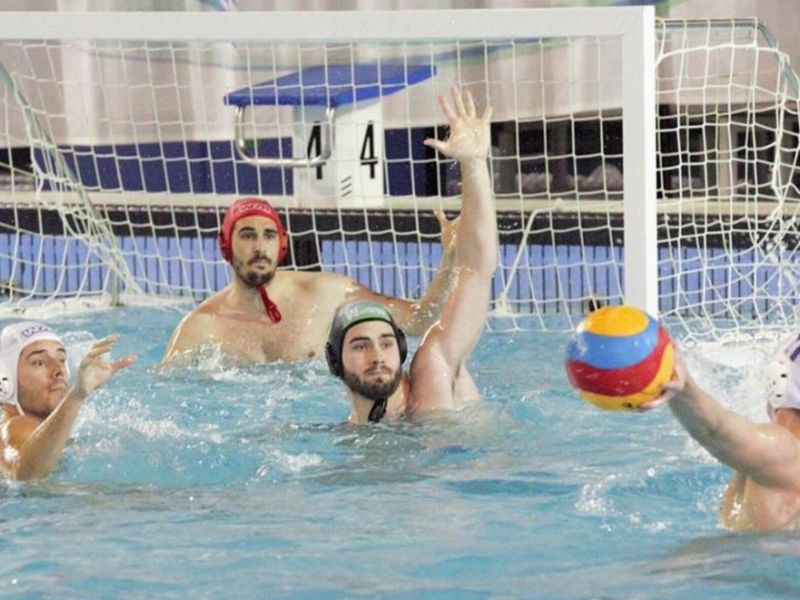 Jugadores de waterpolo en acción durante un partido, con un balón en juego cerca del aro.