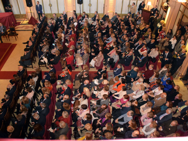 Una multitud de personas se reúne en un auditorio con sillas rojas y tapices dorados. La atmósfera es formal, probablemente en una ceremonia o evento importante.