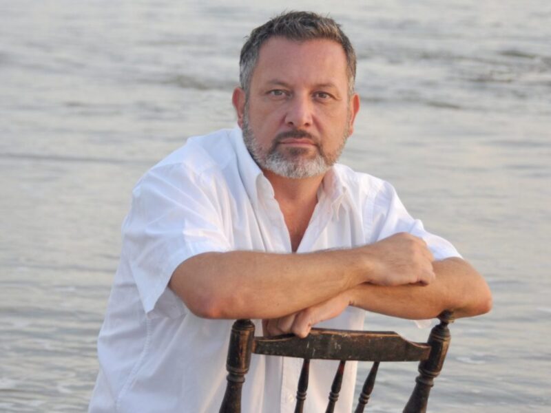 Un hombre con barba gris y cabello corto se sienta en una silla de madera frente al mar. La imagen muestra un paisaje tranquilo con el agua en segundo plano y el hombre mirando directamente a la cámara.