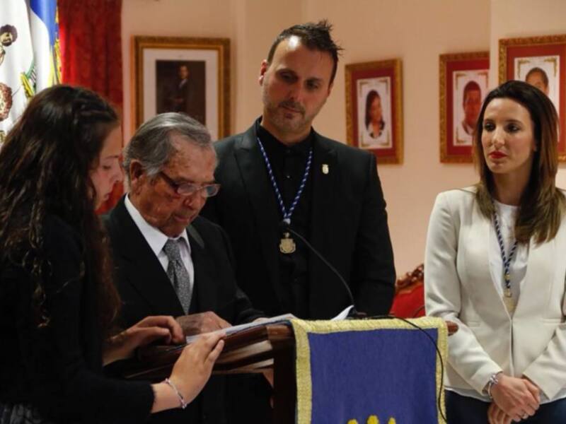Una ceremonia formal con tres personas en el escenario, una mujer en el centro y dos mujeres a su lado, con un fondo de banderas y retratos enmarcados.