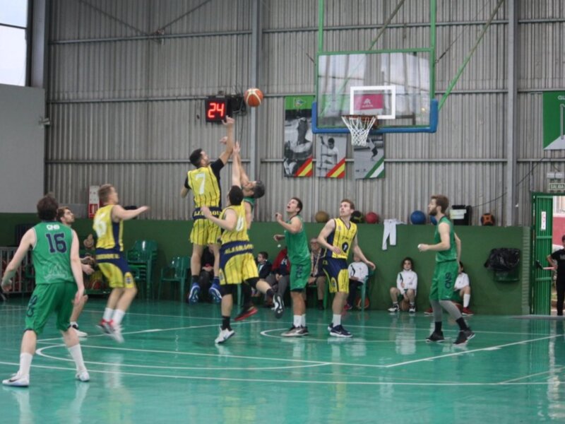 Jugadores en acción durante un partido de baloncesto en una cancha cubierta. Los equipos están identificados con uniformes verdes y amarillos.