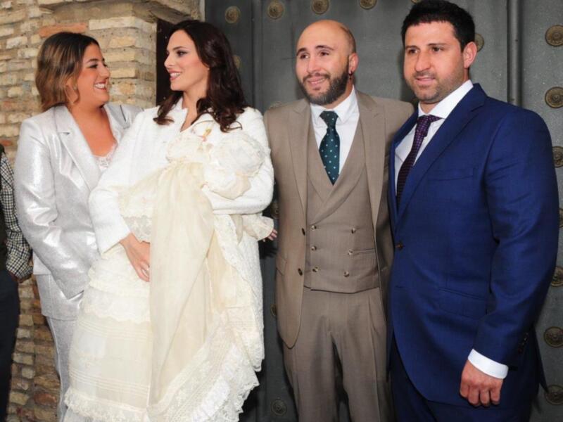 Una familia feliz posa para la foto, con una madre en un vestido blanco, un padre en traje beige y otro hombre en traje azul. La escena parece ser un evento formal o familiar.
