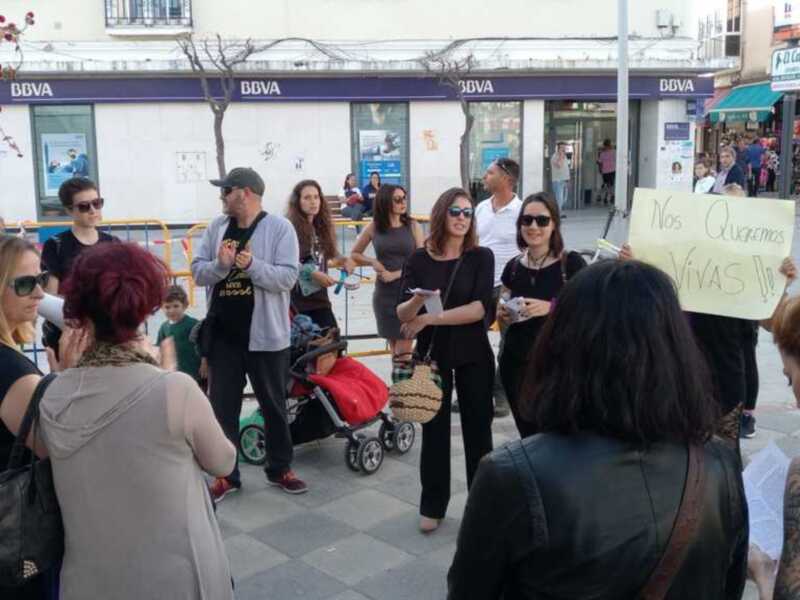 Una multitud de personas se reúne en la calle, algunas con pancartas, frente a una sucursal BBVA. La escena parece ser un evento social o una protesta. En el fondo, se pueden ver edificios y gente caminando. La atmósfera es de concentración y activismo.