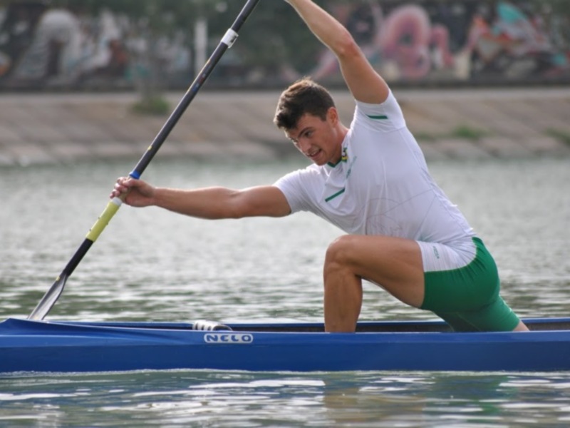 Un atleta de canotaje en acción, con una canoa azul y un fondo de agua y grafitis en el fondo.