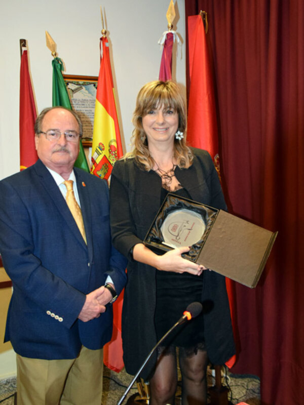 Entrega de un reconocimiento en una ceremonia oficial con banderas y un fondo de cortina roja.