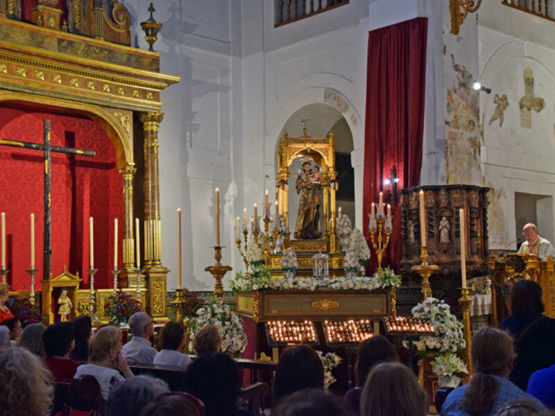 Interior de una iglesia con un altar dorado, velas encendidas y una imagen religiosa en el centro. La decoración incluye flores y una gran cruz en el fondo.