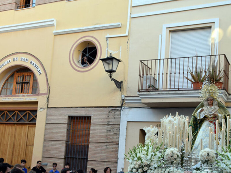 Procesión religiosa frente a la Parroquia de San Basilio, con una imagen de la Virgen María adornada con velas y flores.