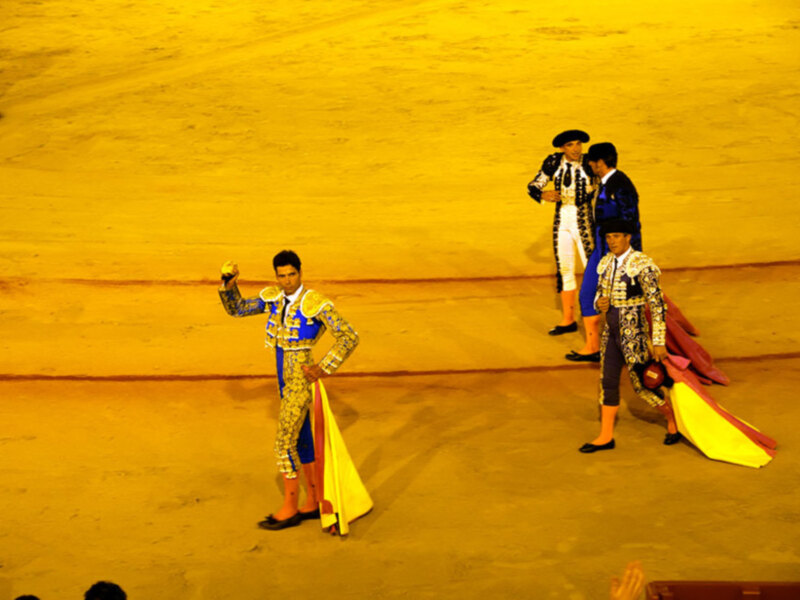 Tres matadores en trajes de luces y mantillas en un ruedo iluminado, preparándose para una corrida de toros.