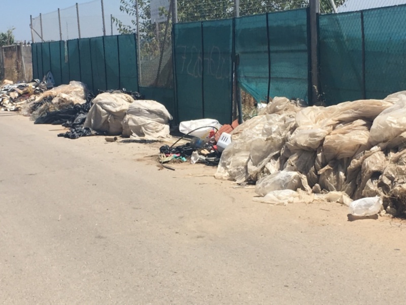 Pila de bolsas de plástico y otros desechos en la calle, cerca de una cerca con una malla de metal y un panel de plástico azul. El paisaje está borroso, lo que sugiere que la imagen se tomó en un entorno urbano con mucha actividad y contaminación.