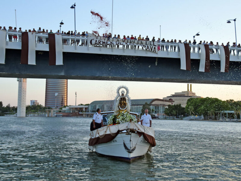 Procesión marítima con la imagen de la Virgen del Carmen en un barco, celebrada bajo un puente con multitud de espectadores.