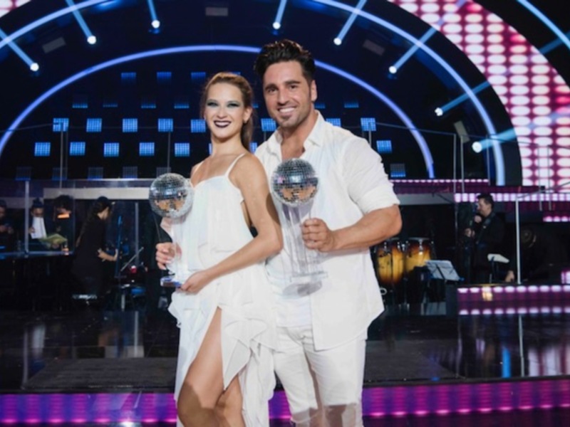 Celebración en escenario con luces y bandas, pareja con trofeos en manos.