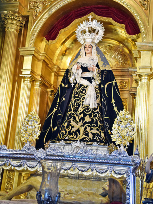 Imaginación de una escena religiosa con una figura vestida de negro y dorado, posiblemente una Virgen María, en un entorno de altar dorado con detalles florales y una figura humana en el fondo. La escena sugiere una representación religiosa o devocional.