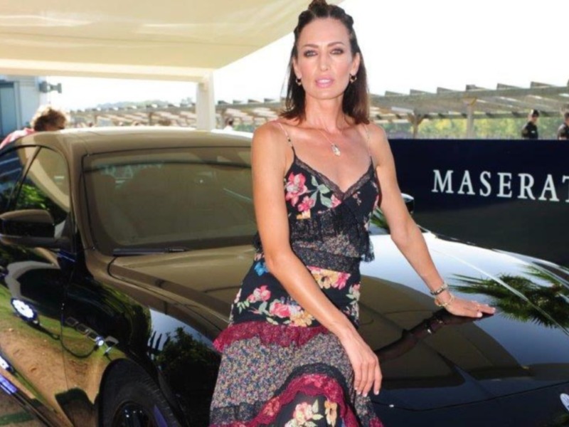 Elegante mujer posando junto a un Maserati en un evento formal. Detalles del vestido floral y la elegancia de la celebridad.