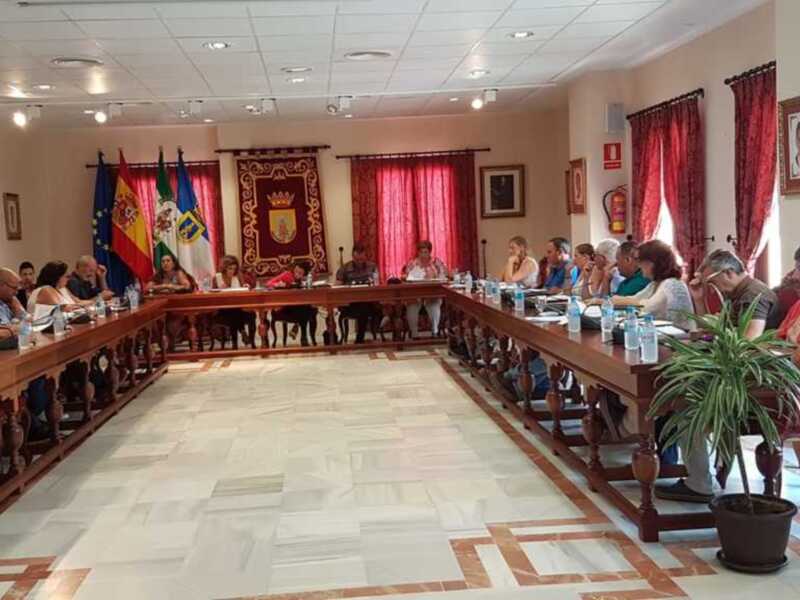 Sala de reuniones con una larga mesa central, personas sentadas en sillas alrededor, banderas y un escudo heráldico en el fondo, iluminación de techo y una planta en el rincón.
