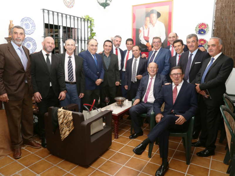 Un grupo de hombres en trajes posa frente a una imagen histórica, probablemente en un evento formal o institucional. La escena está ambientada con una mesa de café y sillas, iluminada por la luz natural que entra a través de un ventanal. El ambiente sugiere una reunión o celebración importante, con los hombres vestidos formalmente para la ocasión.