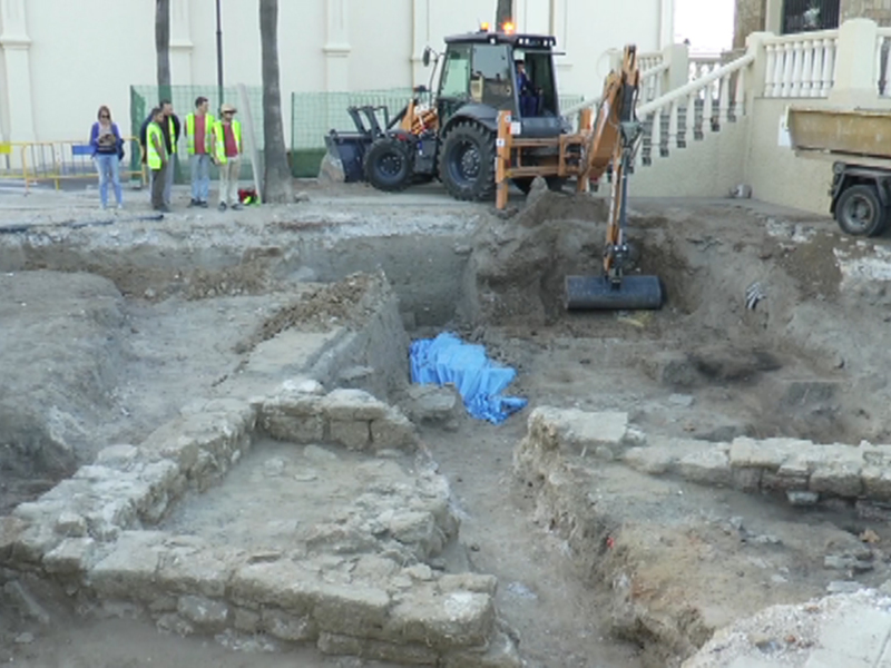 Excavación arqueológica en curso en una zona urbana, con excavadora y equipo de seguridad.