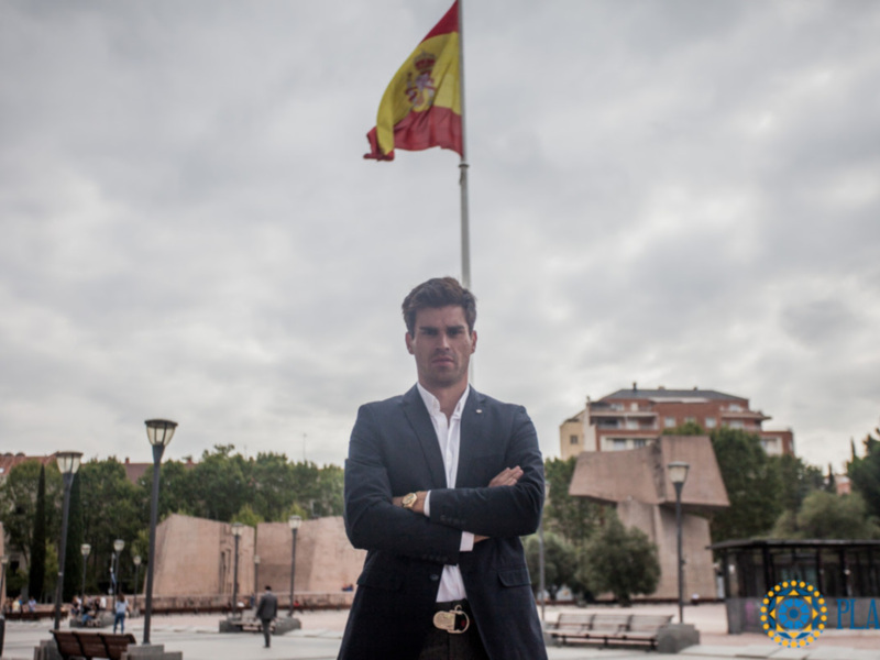 Hombre en traje con brazos cruzados frente a una bandera española en un día nublado.