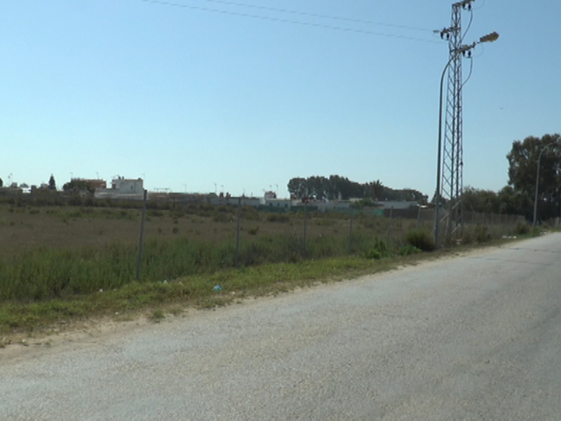 Ruta de acceso a una zona industrial, con campos y edificios en el fondo, bajo un cielo azul claro.