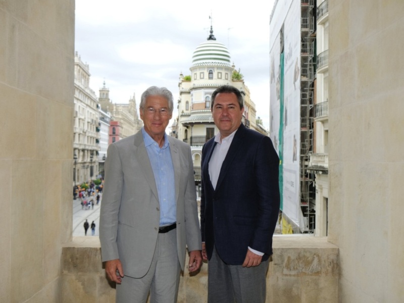 Dos hombres en traje posan juntos en un balcón con vistas a una calle y edificios históricos en el fondo.