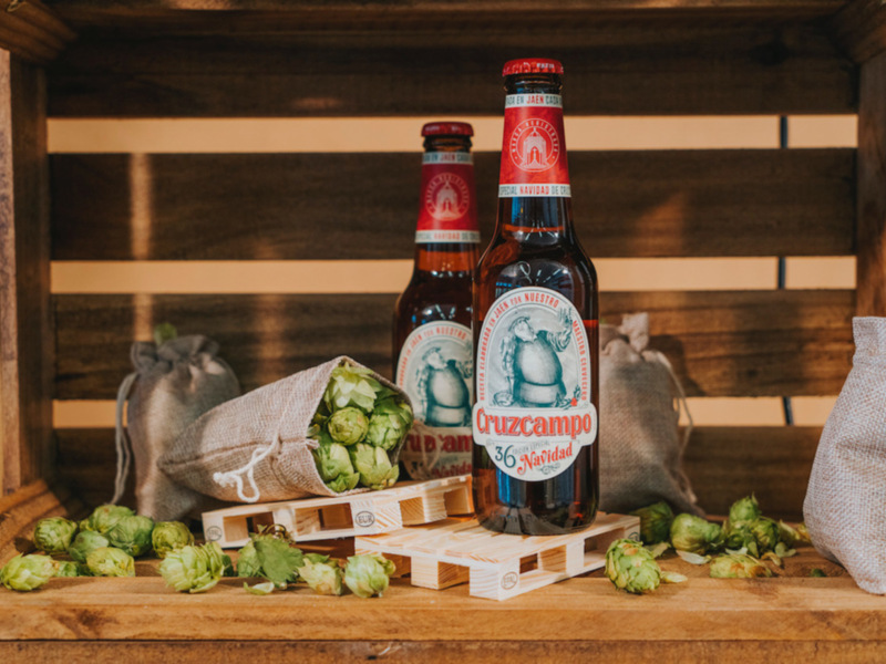 Botellas de cerveza Cruzcampo Navidad en una caja de madera con lúpulo y sacos de lino en el fondo.