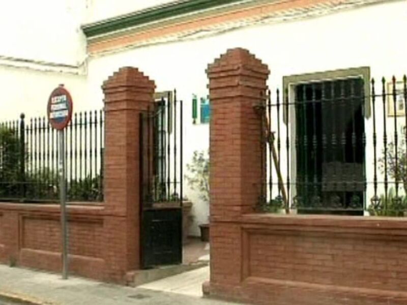 Puerta de acceso a una casa con un diseño elegante, compuesta por una puerta metálica y dos pilares de ladrillo rojo. El entorno incluye un parque con plantas y un cartel de tráfico en el lado izquierdo.