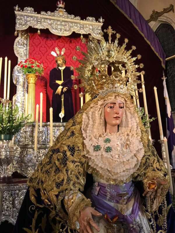 Elegante imagen religiosa con una figura vestida de blanco y dorado, coronada con una grande corona de estrellas y flores, rodeada de velas y una estatua de un personaje en el fondo.