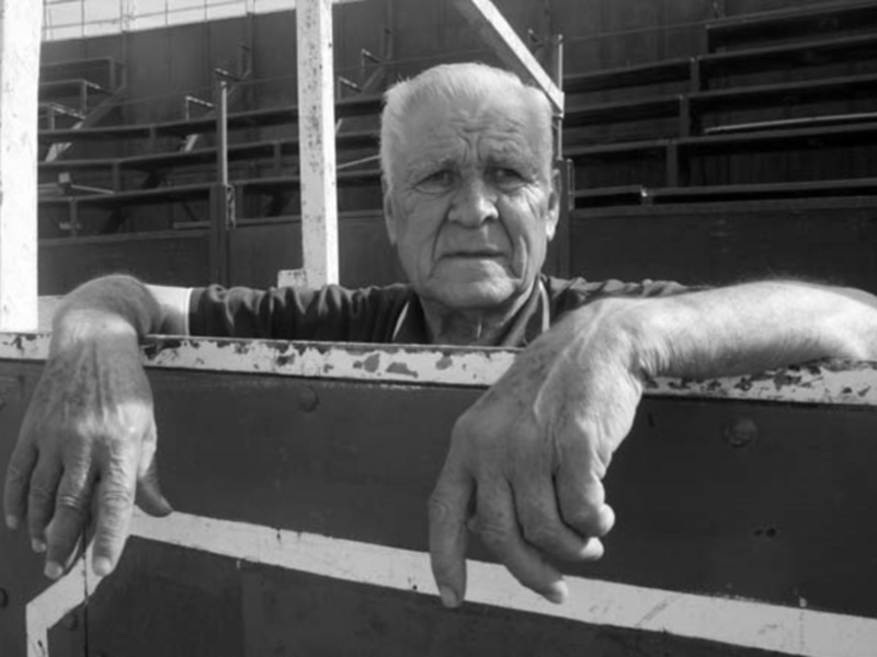 Retrato blanco y negro de un anciano con las manos apoyadas en un marco de madera, en un entorno industrial.